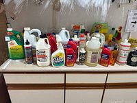 Wide view of approximately 25+ assorted household cleaners on countertop showing varied bottles and labels, some spray tops, some gallon jugs.