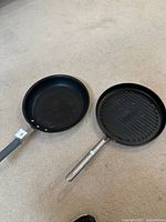 Photo showing a black nonstick skillet with gray handle and a black ridged grill pan with stainless steel handle, both on carpet.