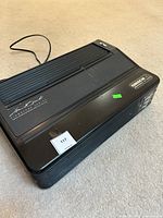 Full view of black Oreck XL Signature Series air purifier unit shown on carpeted floor, showing general form, top surface wear, and power cord extending from back.