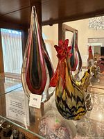 Photo showing the multicolored glass rooster and the tall teardrop-shaped abstract sculpture displayed on a glass shelf with wooden background and reflection.