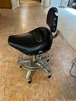 Side angled view showing contoured black saddle seat with rivet detailing on seat and backrest, chrome base with foot ring and rolling casters on tiled floor