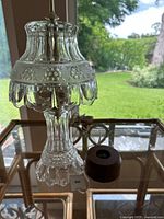 Clear pressed and cut glass vintage electric accent lamp with scalloped floral-etched shade and detailed base, sitting on a glass table with a small round wooden base fixture nearby.