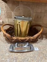 The trio of items consisting of a gold-tone metal ice bucket with a clear handle placed inside a medium oval woven wicker basket on a countertop.