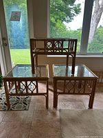 Photo showing set of three vintage rattan tables: two square side tables and one taller rectangular table, all with glass tops and lattice-style side panels, positioned indoors on tiled floor near windows.
