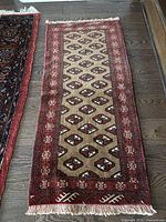 Full view of rectangular Turkmen rug laid on wooden floor showing geometric diamond pattern and red borders with fringed ends