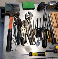 All tools laid out on stainless surface; shows hammer, pliers, wrench, screwdrivers, bit cases, tape measures, boards