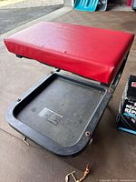 Rolling creeper seat with red cushioned top and black plastic body, shown from above on concrete floor.