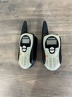 Two Sylvania walkie-talkies side-by-side on a wooden surface showing front with buttons and LCD screens.