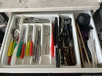 Wide top-down view of white plastic cutlery tray inserted into a drawer holding various plastic-handled and metal cutlery including spoons, forks, and knives, steak knives, scissors, and chopsticks.