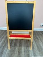 Front view of the wooden kids art stand with black chalkboard surface and red storage tray below.