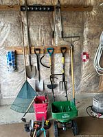 Full view of large gardening tools including rakes, shovels, grass hook, sprinkler, lawn spreader, and red buckets with hand tools.