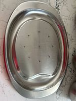 Oval metal meat tray with multiple drainage holes on a marble countertop