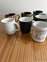 Seven assorted ceramic coffee mugs displayed on wooden surface, showing varied patterns including polka dots and cartoon graphics.