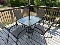 Four sling fabric chairs around square outdoor patio table with glass top on a wooden deck.