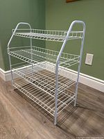 Front side view of white metal shoe rack with four wire shelves on wood floor against green wall.
