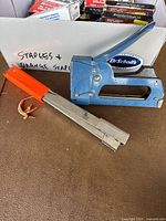 Photo of two staplers and assorted boxes of staples in white container with label Staples + Orange Staples