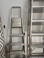 Both aluminum step ladders side by side against a wall showing height and step count comparison.
