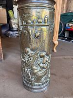 Full view of brass stick stand showing embossed scene of two people, one playing guitar, seated at a table with kitchenware and pottery on shelves above