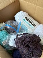 Top-down view of a cardboard box containing multiple skeins of yarn including a large white skein Bernat Handicrafter cotton acrylic blend and other assorted colors like navy, teal, and brown.