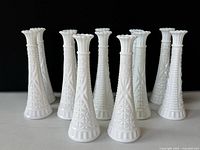 10 white milk glass bud vases arranged in grouped sets against black background, showing detailed texture and shape.