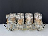 Front view of eight decorated highball glasses in the metal wire caddy showing gold leaf and frosted starburst pattern