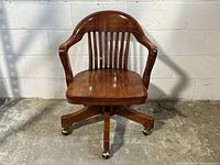 Front view of antique style Bombay Company bankers chair showing curved backrest with vertical slats, wooden arms and brass-retreaded caster wheels on a five-legged wooden base.
