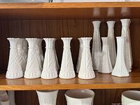 Front view of nine vintage white milk glass bud vases displayed on a wooden shelf, showing varying shapes and embossed floral and geometric patterns.