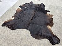 Full view of the dark brown cowhide rug laid flat on a light gray carpet showing natural shape and texture.