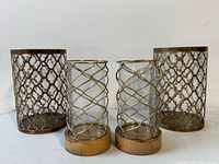 Four decorative candle holders: two large glass holders with brown lattice metal overlay and two smaller glass holders with gold spiral metal design on wooden bases.