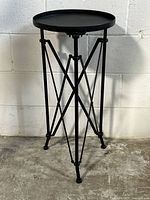 Black metal round top side table shown from a front left angle, highlighting the industrial metal leg design and round tray style top.