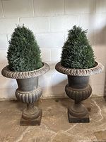 Both urn planters with faux cedar shrubs side by side against wall on concrete floor.