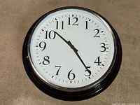 Full front view of the large round wall clock lying flat showing the black frame with silver chrome rim and white dial with black numbers and hands.