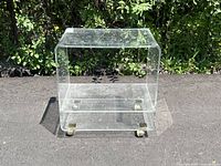 Front view of clear lucite end table with brass-toned caster wheels on a paved surface.