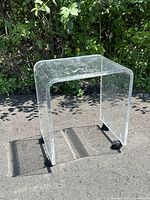 Clear lucite waterfall end table on black rollers shown outdoors with sunlight and shadows highlighting the transparent material and scratches.
