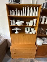 Frontal view of vintage mid-century modern teak wall unit with three shelves filled with white milk glass collectibles, drop down cabinet door, and three drawers below.
