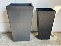 Front view of two black tapered plastic planters side by side on floor against a white wall, showing size difference and surface wear.