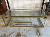 Front view of rectangular brass tone metal frame coffee table with glass top and lower shelf, showing the geometric design and clear surfaces.