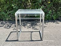 Front and side view of clear lucite side table with shiny chrome base placed outdoors with natural lighting.