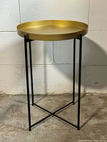 Full view of the metal tray table showing the gold tray top on the black iron base against a concrete floor and white wall background.
