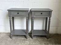 Two matching grey bedside tables with one drawer and lower shelf each, shown side by side against a white wall on concrete floor. Clear view of the tables' front and sides, showcasing the built-in electrical and USB ports on the side of each table.