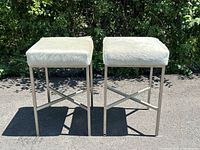 Pair of modern barstools shown outdoors on pavement in front of greenery. Each barstool has brushed metal legs and a square natural cream cowhide upholstered seat.