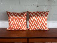 Two orange and cream zigzag patterned square pillows with button closure backs placed side by side on a wood surface against a white wall, showing minor fabric pulling and mild wear.