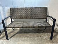 Front view of black metal frame outdoor bench with resin wicker seat and backrest against a white brick wall on concrete floor.