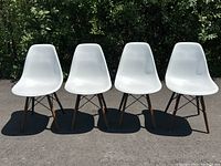 Four white molded fiberglass seat chairs with dark brown wood legs shown outdoors on pavement in front of greenery.