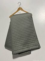 Quilted grey coverlet hanging on a wooden hanger showing square stitched pattern and border on light backdrop.