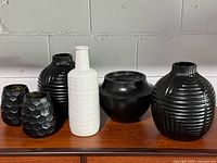 Six ceramic vases including five black and one white vase arranged side by side on wooden surface against a white wall. Variety of shapes and textures visible in photo.