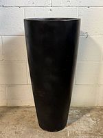 Full front view of tall black cylindrical resin planter on concrete floor against white brick wall.