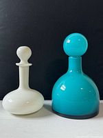 Two Mid-Century Modern style cased glass decanters shown together, one turquoise and one white with rounded stoppers, placed against a black background.
