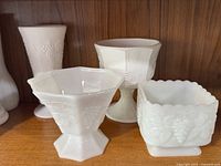 Four pieces of white milk glass arranged on wooden shelf, showing variety of shapes and the Harvest grape embossed pattern.