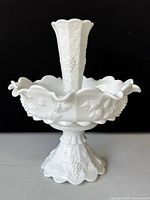 Front view of the vintage milk glass epergne centerpiece showing the grape and leaf pattern on the bowl, central trumpet vase, and pedestal base, all in white opaque milk glass with scalloped edges.